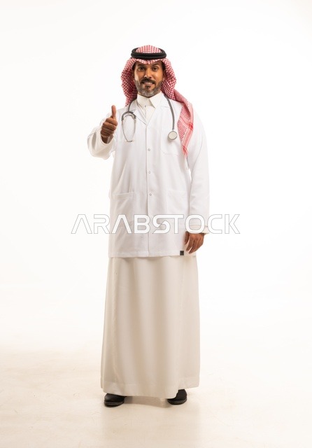 أشارة أصبع الأبهام للأعلى النظر إلى الكاميرا بايماءات الثقة بالنفس، الاهتمام بالمظهر الخارجي، تقديم الفحص الطبي والخدمات العلاجية، بورتريه مقرب لطبيب عربي خليجي سعودي مبتسم يرتدي معطف الطبيب ويضع السماعات الطبية، مفهوم الرعاية الصحية، خلفية بيضاء