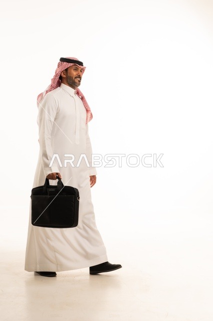 مفهوم إداة الأعمال والمشاريع، الاستعداد للذهاب إلى العمل، بورتريه لرجل عربي خليجي سعودي يرتدي الثوب التقليدي والشماغ يحمل بيده حقيبة يد يقف باستقامة بإيماءات الثقة بالنفس، الأناقة والاهتمام بالمظهر الخارجي، الجسم بالطول الكامل، خلفيه بيضاء