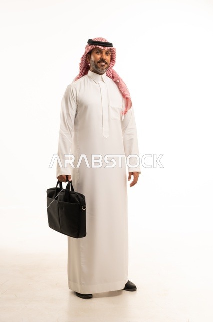 مفهوم إداة الأعمال والمشاريع، الاستعداد للذهاب إلى العمل، بورتريه لرجل عربي خليجي سعودي يرتدي الثوب التقليدي والشماغ يحمل بيده حقيبة يد يقف باستقامة بإيماءات الثقة بالنفس، الأناقة والاهتمام بالمظهر الخارجي، الجسم بالطول الكامل، خلفيه بيضاء