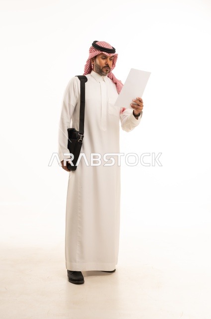 قراءة وتدقيق الملاحظات، مفهوم إداة الأعمال والمشاريع، الاستعداد للذهاب إلى العمل، بورتريه لرجل عربي خليجي سعودي يرتدي الثوب التقليدي والشماغ يحمل بيده حقيبة يد وورقة بيضاء، الأناقة والاهتمام بالمظهر الخارجي، الجسم بالطول الكامل، خلفيه بيضاء