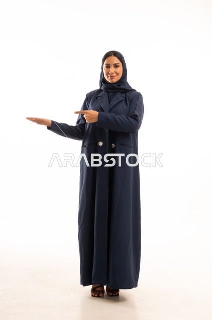أشارة يدين تفضل الوجهة من هنا، الاهتمام بالمظهر الخارجي والأناقة، تعابير سرور وابتسامة ، بورتريه لإمرأة عربية خليجية سعودية محجبة ترتدي عباءة، شابة سعودية ،مفهوم التعبير بالإشارة، صورة الجسم بالطول الكامل، خلفية بيضاء