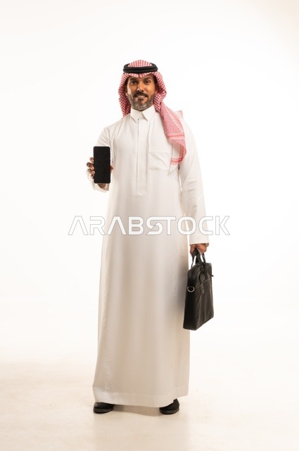 استخدام جهاز تقني حديث ومتطور، مفهوم إداة الأعمال والمشاريع، الاستعداد للذهاب إلى العمل، بورتريه لرجل عربي خليجي سعودي يرتدي الثوب التقليدي والشماغ يحمل بيده حقيبة يد وورقة بيضاء ويقوم باستخدام الهاتف المحمول، الجسم بالطول الكامل، خلفيه بيضاء