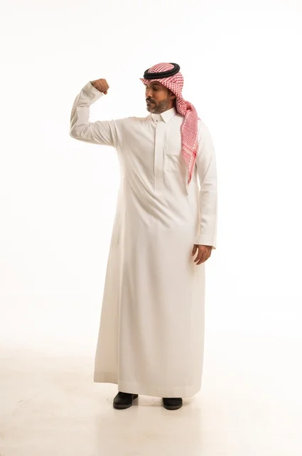 Increasing muscle endurance, mastering boxing techniques, a portrait of an Arab Gulf Saudi man wearing traditional attire raising his fists in gestures of strength, looking at the camera with expressions of self-confidence, practicing martial arts, full-body shot, white background.