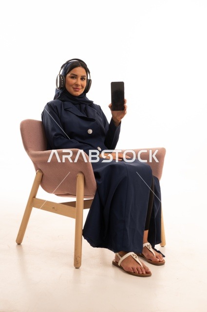 عرض شاشة سوداء على الهاتف المحمول، استخدام الهاتف المحمول في الحياة اليومية، الاهتمام بالمظهر الخارجي والأناقة، تعابير الابتسامة والسرور، بورتريه لإمرأة عربية خليجية سعودية محجبة ترتدي عباءة تجلس على كرسي وردي وتضع سماعة الأذن، شابة سعودية ، خلفية بيضاء