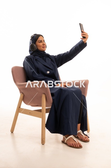 اجراء مكالمة هاتفية، استخدام الهاتف المحمول في الحياة اليومية، الاهتمام بالمظهر الخارجي والأناقة، تعابير الابتسامة والسرور، بورتريه لإمرأة عربية خليجية سعودية محجبة ترتدي عباءة تجلس على كرسي وردي ، شابة سعودية ، خلفية بيضاء
