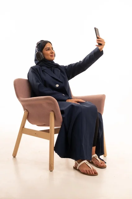 اجراء مكالمة هاتفية، استخدام الهاتف المحمول في الحياة اليومية، الاهتمام بالمظهر الخارجي والأناقة، تعابير الابتسامة والسرور، بورتريه لإمرأة عربية خليجية سعودية محجبة ترتدي عباءة تجلس على كرسي وردي ، شابة سعودية ، خلفية بيضاء