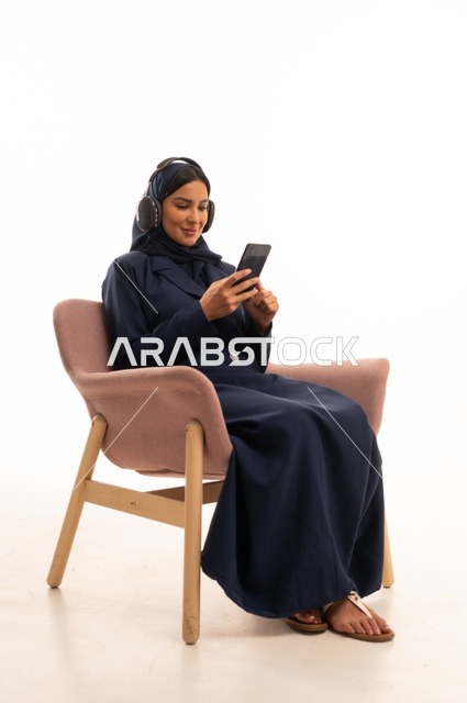 استخدام الهاتف المحمول في الحياة اليومية، الاهتمام بالمظهر الخارجي والأناقة، تعابير الابتسامة والسرور، بورتريه لإمرأة عربية خليجية سعودية محجبة ترتدي عباءة تجلس على كرسي وردي ، شابة سعودية تتصفح مواقع التواصل الاجتماعي، خلفية بيضاء