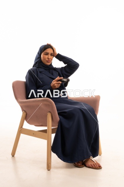 Hand and face expressions, hand on the head indicate loss, concern for appearance and elegance, expressions of sadness and regret, a portrait of a Saudi Arabian Gulf woman wearing a hijab and an abaya sitting on a pink chair holding a fist, a young Saudi woman playing video games, white background.