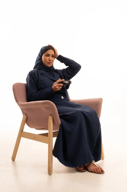Hand and face expressions, hand on the head indicate loss, concern for appearance and elegance, expressions of sadness and regret, a portrait of a Saudi Arabian Gulf woman wearing a hijab and an abaya sitting on a pink chair holding a fist, a young Saudi woman playing video games, white background.