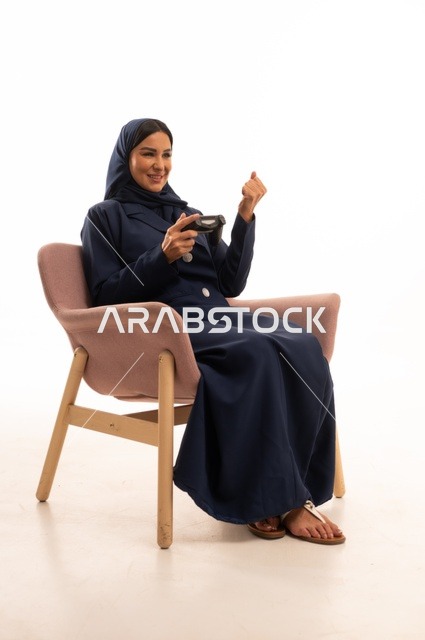 تعابير يد ووجه قبضة يد تدل على الفوز والانتصار، الاهتمام بالمظهر الخارجي والأناقة، تعابير السرور والسعادة، بورتريه لإمرأة عربية خليجية سعودية محجبة ترتدي عباءة تجلس على كرسي وردي تمسك قبضة يد، شابة سعودية تلعب ألعاب الفيديو، خلفية بيضاء