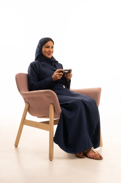 قضاء أوقات ترفيهية ممتعة، الاهتمام بالمظهر الخارجي والأناقة، تعابير السرور والسعادة، بورتريه لإمرأة عربية خليجية سعودية محجبة ترتدي عباءة تجلس على كرسي وردي زتمسك قبضة يد، شابة سعودية تلعب العاب الفيديو، خلفية بيضاء