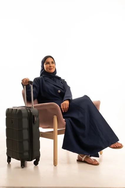 النظر إلى الأعلى بإيماءات التمني والتأمل، الاهتمام بالمظهر الخارجي والأناقة، بورتريه لإمرأة عربية خليجية سعودية محجبة ترتدي عباءة تجلس على كرسي وردي وتمسك بيدها حقيبة سفر سوداء، شابة سعودية ، خلفية بيضاء