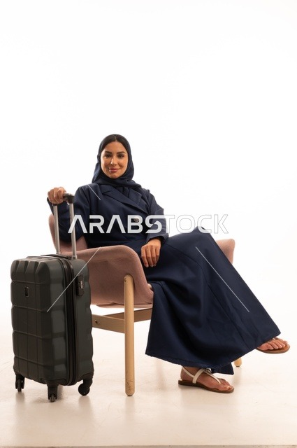 النظر إلى الكاميرا بإيماءات السرور السعادة، الاهتمام بالمظهر الخارجي والأناقة، بورتريه لإمرأة عربية خليجية سعودية محجبة ترتدي عباءة تجلس على كرسي وردي وتمسك بيدها حقيبة سفر سوداء، شابة سعودية ، خلفية بيضاء