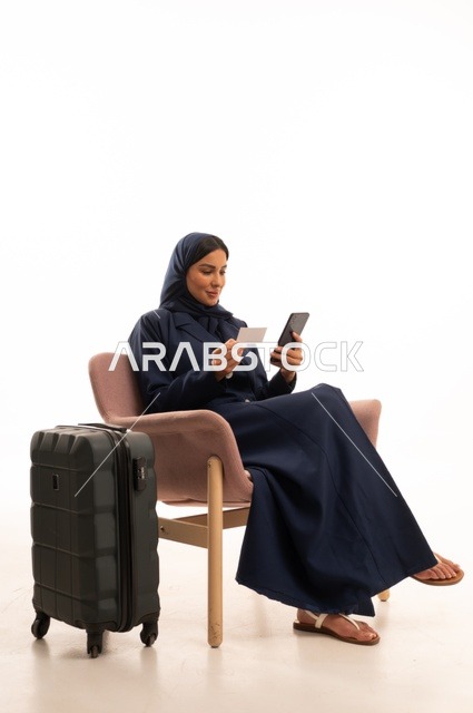 مطابقة بيانات الرحلة الشخصية مع الرحلات الأخرى، دمج التقنية الحديثة في الحياة اليومية الاهتمام بالمظهر الخارجي والأناقة، بورتريه لإمرأة عربية خليجية سعودية محجبة ترتدي عباءة تجلس على كرسي وردي وبجانبها حقيبة سفر سوداء، شابة سعودية ، خلفية بيضاء