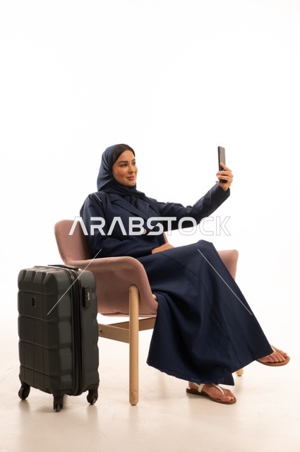 التقاط صورة سيلفي من خلال جهاز الهاتف للاحتفاظ بالذكريات، دمج التقنية الحديثة في الحياة اليومية الاهتمام بالمظهر الخارجي والأناقة، بورتريه لإمرأة عربية خليجية سعودية محجبة ترتدي عباءة تجلس على كرسي وردي وبجانبها حقيبة سفر سوداء، شابة سعودية ، خلفية بيضاء