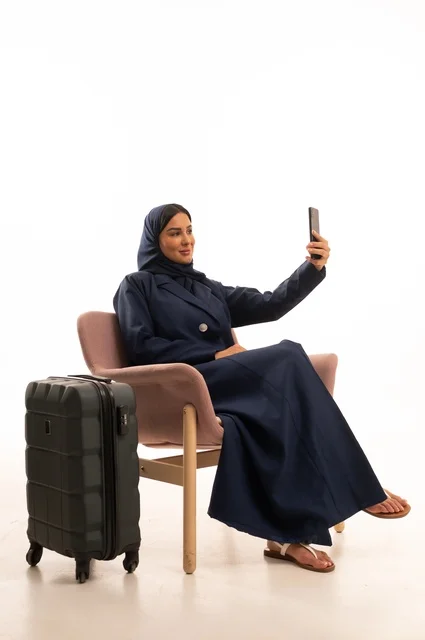 التقاط صورة سيلفي من خلال جهاز الهاتف للاحتفاظ بالذكريات، دمج التقنية الحديثة في الحياة اليومية الاهتمام بالمظهر الخارجي والأناقة، بورتريه لإمرأة عربية خليجية سعودية محجبة ترتدي عباءة تجلس على كرسي وردي وبجانبها حقيبة سفر سوداء، شابة سعودية ، خلفية بيضاء