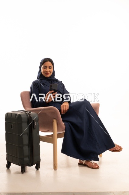 متابعة الرحلات الجوية من خلال جهاز الهاتف المحمول، دمج التقنية الحديثة في الحياة اليومية الاهتمام بالمظهر الخارجي والأناقة، بورتريه لإمرأة عربية خليجية سعودية محجبة ترتدي عباءة تجلس على كرسي وردي وبجانبها حقيبة سفر سوداء، شابة سعودية ، خلفية بيضاء