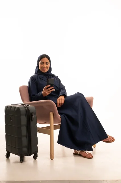 متابعة الرحلات الجوية من خلال جهاز الهاتف المحمول، دمج التقنية الحديثة في الحياة اليومية الاهتمام بالمظهر الخارجي والأناقة، بورتريه لإمرأة عربية خليجية سعودية محجبة ترتدي عباءة تجلس على كرسي وردي وبجانبها حقيبة سفر سوداء، شابة سعودية ، خلفية بيضاء