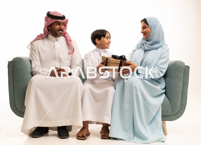 الألفة والمودة بين أفراد الأسرة السعودية, بورتريه  لعائلة عربية خليجية سعودية تجلس على الاريكة تقدم الهدية لولدها بايماءات الرضا والسرور, تقديم الهدايا بالمناسبات وأعياد الميلاد, خلفية بيضاء