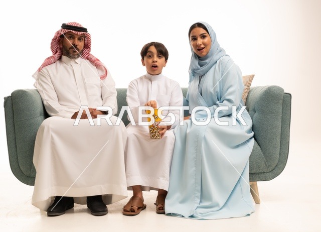 Happy family atmosphere, sharing snacks with the family, a portrait of a Saudi Gulf Arab family sitting on a comfortable couch watching television, watching movies and series, recreational weekend activities, enjoying eating popcorn, white background.