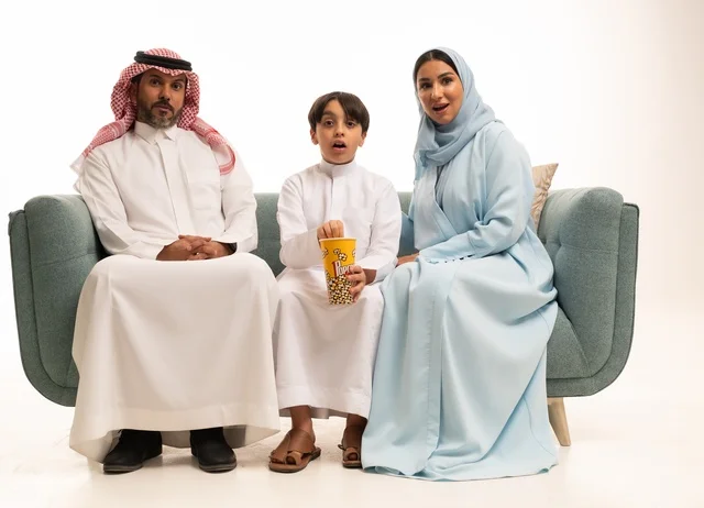أجواء عائلية سعيدة، مشاركة الوجبات الخفيفة مع العائلة، بورتريه لأسرة عربية خليجية سعودية يجلسون على كنبة مريحة يقومون بمتابعة التلفاز، مشاهدة الافلام والمسلسلات، أنشطة عطلة نهاية الاسبوع الترفيهية، الاستمتاع بتناول الفوشار، خلفية بيضاء
