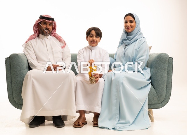 أجواء عائلية سعيدة، مشاركة الوجبات الخفيفة مع العائلة، بورتريه لأسرة عربية خليجية سعودية يجلسون على كنبة مريحة يقومون بمتابعة التلفاز، مشاهدة الافلام والمسلسلات، أنشطة عطلة نهاية الاسبوع الترفيهية، الاستمتاع بتناول الفوشار، خلفية بيضاء