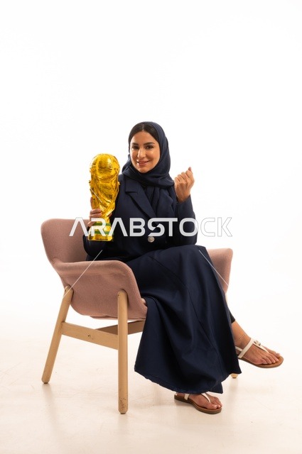 ايماءات الفوز والنصر، كأس العالم ذهبي لامع،الاهتمام بالمظهر الخارجي والأناقة، بورتريه لإمرأة عربية خليجية سعودية محجبة ترتدي عباءة تجلس على كرسي وردي تمسك بيدها كأس الفوز في المباراة، شابة سعودية ، خلفية بيضاء