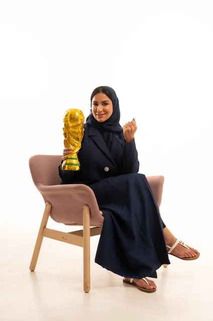 ايماءات الفوز والنصر، كأس العالم ذهبي لامع،الاهتمام بالمظهر الخارجي والأناقة، بورتريه لإمرأة عربية خليجية سعودية محجبة ترتدي عباءة تجلس على كرسي وردي تمسك بيدها كأس الفوز في المباراة، شابة سعودية ، خلفية بيضاء