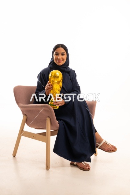 ايماءات الفوز والنصر، كأس العالم ذهبي لامع،الاهتمام بالمظهر الخارجي والأناقة، بورتريه لإمرأة عربية خليجية سعودية محجبة ترتدي عباءة تجلس على كرسي وردي تمسك بيدها كأس الفوز في المباراة، شابة سعودية ، خلفية بيضاء