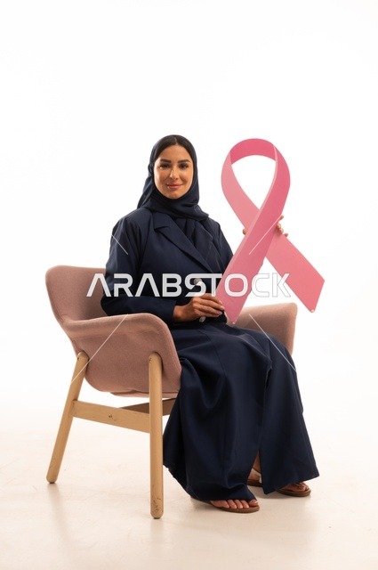 التوعية الصحية التوعية بالفحص المبكر لسرطان الثدي، الاهتمام بالمظهر الخارجي والأناقة، بورتريه لإمرأة عربية خليجية سعودية محجبة ترتدي عباءة تجلس على كرسي وردي تمسك بيدها الاشارة الوردية، أهمية الفحوصات الدورية، شهر أكتوبر، شابة سعودية ، خلفية بيضاء
