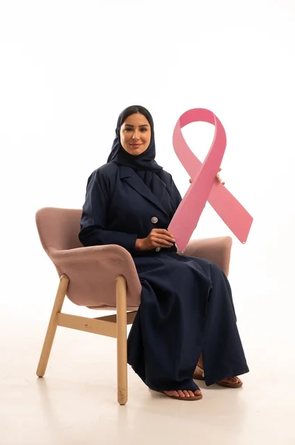 التوعية الصحية التوعية بالفحص المبكر لسرطان الثدي، الاهتمام بالمظهر الخارجي والأناقة، بورتريه لإمرأة عربية خليجية سعودية محجبة ترتدي عباءة تجلس على كرسي وردي تمسك بيدها الاشارة الوردية، أهمية الفحوصات الدورية، شهر أكتوبر، شابة سعودية ، خلفية بيضاء