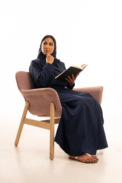تعابير التفكير والتخيل، المطالعة والقراءة اكتساب معلومات ثقافية جديدة،الاهتمام بالمظهر الخارجي والأناقة، بورتريه لإمرأة عربية خليجية سعودية محجبة ترتدي عباءة تجلس على كرسي وردي تمسك بيدها كتاب، شابة سعودية ، خلفية بيضاء