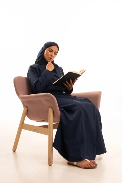 تعابير الاندماج والتركيز، المطالعة والقراءة اكتساب معلومات ثقافية جديدة،الاهتمام بالمظهر الخارجي والأناقة، بورتريه لإمرأة عربية خليجية سعودية محجبة ترتدي عباءة تجلس على كرسي وردي تمسك بيدها كتاب، شابة سعودية ، خلفية بيضاء