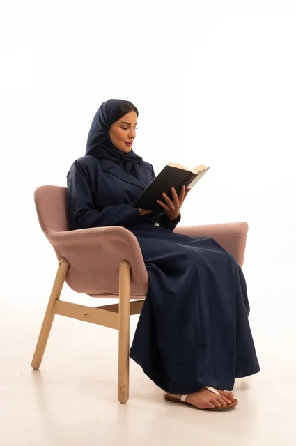 تعابير الاندماج والتركيز، المطالعة والقراءة اكتساب معلومات ثقافية جديدة،الاهتمام بالمظهر الخارجي والأناقة، بورتريه لإمرأة عربية خليجية سعودية محجبة ترتدي عباءة تجلس على كرسي وردي تمسك بيدها كتاب، شابة سعودية ، خلفية بيضاء