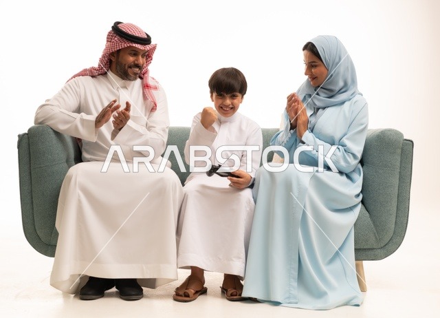 The concept of a happy family, a portrait of a Saudi Gulf Arab family sitting in the living room, enjoying their time playing video games with gestures indicating happiness, a boy holding a controller and playing video games next to his parents, fun and entertainment with recreational PlayStation games, white background.