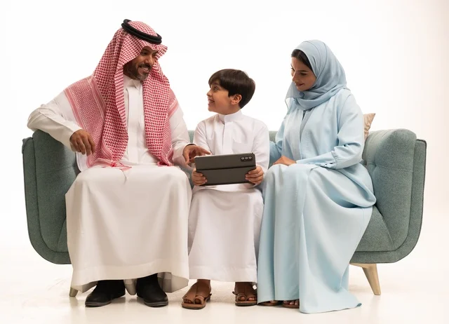 استخدام جهاز تقني حديث، متابعة حصص الأونلاين وإجراء مكالمة فيديو من خلال الجهاز اللوحي، بورتريه لولد عربي سعودي خليجي يرتدي الثوب التقليدي يحمل التابليت ويجلس على الاريكة برفقة والديه، الأجواء العائلية المليئة بالحب والترابط والإستقرار، خلفية بيضاء