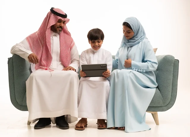 Using a modern technical device, attending online classes and making a video call through the tablet, a portrait of an Arab Saudi Gulf boy wearing traditional attire holding a tablet and sitting on the sofa with his parents, a family atmosphere filled with love, connection, and stability, white background.