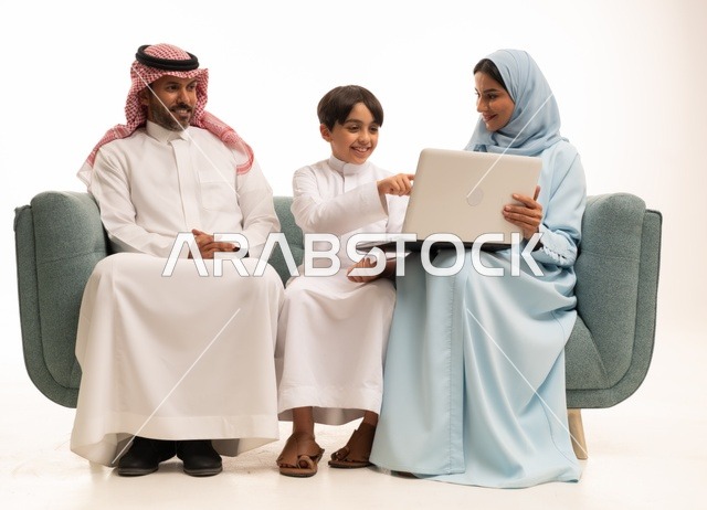 متابعة حصص الأونلاين وإجراء مكالمة فيديو من خلال الحاسوب المحمول، استخدام جهاز تقني حديث، بورتريه لولد عربي سعودي خليجي يرتدي الثوب التقليدي يحمل الحاسوب ويجلس على الاريكة برفقة والديه، الأجواء العائلية المليئة بالحب والترابط والإستقرار، خلفية بيضاء