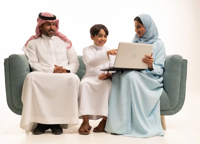 متابعة حصص الأونلاين وإجراء مكالمة فيديو من خلال الحاسوب المحمول، استخدام جهاز تقني حديث، بورتريه لولد عربي سعودي خليجي يرتدي الثوب التقليدي يحمل الحاسوب ويجلس على الاريكة برفقة والديه، الأجواء العائلية المليئة بالحب والترابط والإستقرار، خلفية بيضاء
