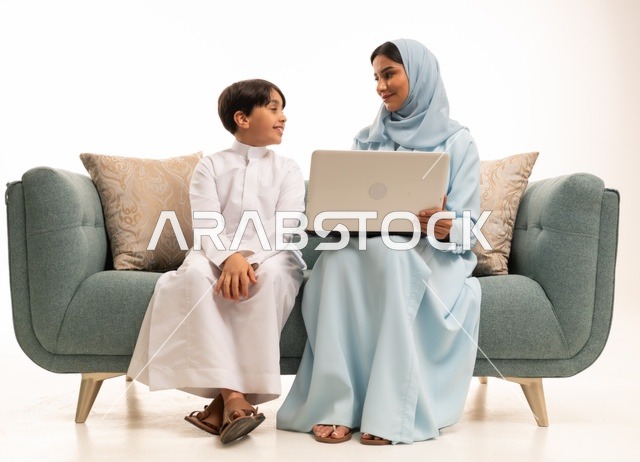Following online classes and making a video call through a laptop, using modern technology, a portrait of an Arab Saudi Gulf boy wearing traditional attire using a computer and sitting on the couch with his mother, a family atmosphere filled with love, connection, and stability, white background.