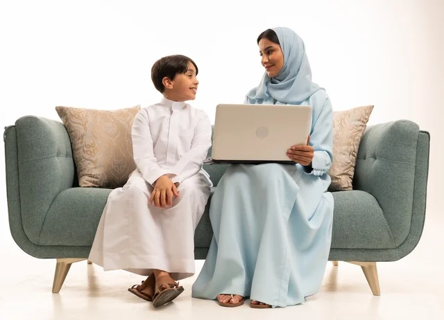 متابعة حصص الأونلاين وإجراء مكالمة فيديو من خلال الحاسوب المحمول، استخدام جهاز تقني حديث، بورتريه لولد عربي سعودي خليجي يرتدي الثوب التقليدي يستخدم الحاسوب ويجلس على الاريكة برفقة والدته، الأجواء العائلية المليئة بالحب والترابط والاستقرار، خلفية بيضاء