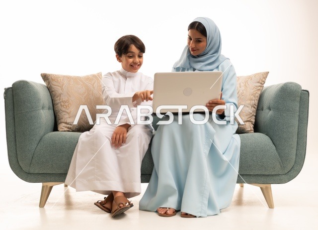 Following online classes and making a video call through a laptop, using modern technology, a portrait of an Arab Saudi Gulf boy wearing traditional attire using a computer and sitting on the couch with his mother, a family atmosphere filled with love, connection, and stability, white background.