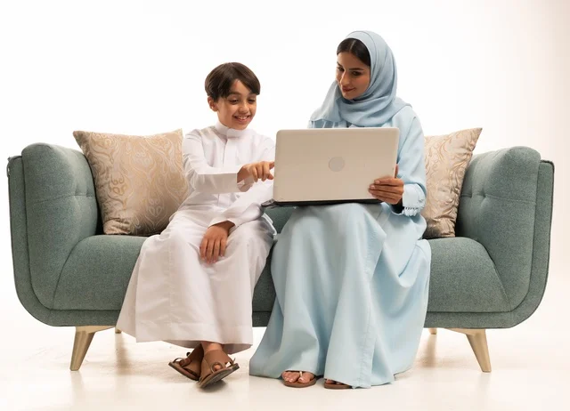 متابعة حصص الأونلاين وإجراء مكالمة فيديو من خلال الحاسوب المحمول، استخدام جهاز تقني حديث، بورتريه لولد عربي سعودي خليجي يرتدي الثوب التقليدي يستخدم الحاسوب ويجلس على الاريكة برفقة والدته، الأجواء العائلية المليئة بالحب والترابط والاستقرار، خلفية بيضاء