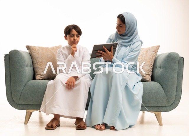 The family atmosphere filled with love, connection, and stability, attending online classes and making a video call through a mobile tablet, using modern technology, a portrait of an Arab Saudi Gulf boy wearing traditional attire using the tablet and sitting on the sofa with his mother, white background.