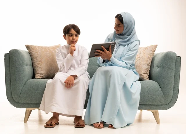 الأجواء العائلية المليئة بالحب والترابط والاستقرار، متابعة حصص الأونلاين وإجراء مكالمة فيديو من خلال التابليت المحمول، استخدام جهاز تقني حديث، بورتريه لولد عربي سعودي خليجي يرتدي الثوب التقليدي يستخدم الجهاز اللوحي ويجلس على الاريكة برفقة والدته، خلفية بيضاء