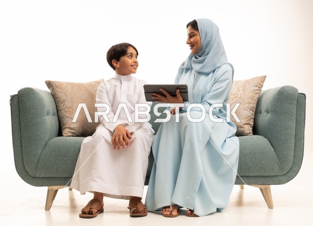 الأجواء العائلية المليئة بالحب والترابط والاستقرار، متابعة حصص الأونلاين وإجراء مكالمة فيديو من خلال التابليت المحمول، استخدام جهاز تقني حديث، بورتريه لولد عربي سعودي خليجي يرتدي الثوب التقليدي يستخدم الجهاز اللوحي ويجلس على الاريكة برفقة والدته، خلفية بيضاء