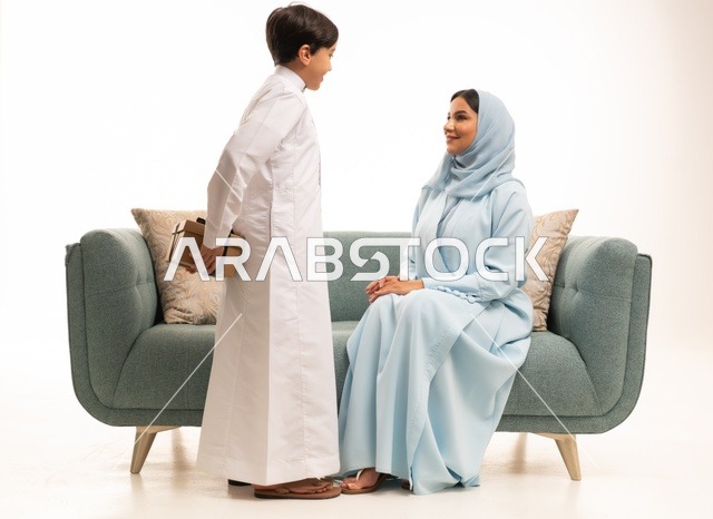 مشاعر الحب والحنان بمنح الأم هدايا العيد، تقدير الوالدين ومفهوم الإهداء, أجواء لمة الأهل في المناسبات السعيدة, بورتريه لولد عربي خليجي سعودي يرتدي الثوب التقليدي يقوم بإعطاء أمه هدية بإيماءات السعادة, عائلة سعودية تحتفل بالعيد المبارك, خلفية بيضاء