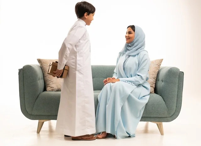 Feelings of love and tenderness in giving the mother Eid gifts, appreciation for parents and the concept of gifting, the atmosphere of family gatherings during happy occasions, a portrait of an Arab Gulf Saudi boy wearing traditional attire giving his mother a gift with gestures of happiness, a Saudi family celebrating the blessed Eid, white background.