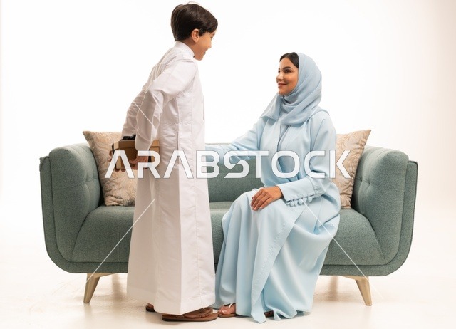 مشاعر الحب والحنان بمنح الأم هدايا العيد، تقدير الوالدين ومفهوم الإهداء, أجواء لمة الأهل في المناسبات السعيدة, بورتريه لولد عربي خليجي سعودي يرتدي الثوب التقليدي يقوم بإعطاء أمه هدية بإيماءات السعادة, عائلة سعودية تحتفل بالعيد المبارك, خلفية بيضاء