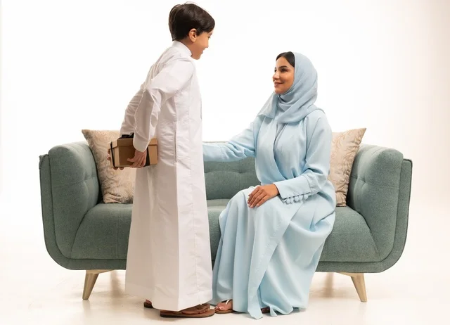 مشاعر الحب والحنان بمنح الأم هدايا العيد، تقدير الوالدين ومفهوم الإهداء, أجواء لمة الأهل في المناسبات السعيدة, بورتريه لولد عربي خليجي سعودي يرتدي الثوب التقليدي يقوم بإعطاء أمه هدية بإيماءات السعادة, عائلة سعودية تحتفل بالعيد المبارك, خلفية بيضاء